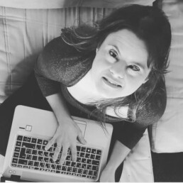 #Audiodescrição: Foto em preto e branco. Mulher branca com Síndrome de Down. Tem cabelo longo, está com um laptop no colo e olha para cima.