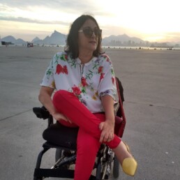 #Audiodescrição: Mulher branca em cadeira de rodas. Ela tem cabelo preto na altura dos ombros. Usa óculos escuros, camisa branca com flores vermelhas e calça vermelha.