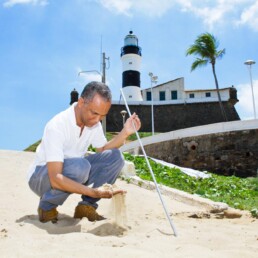 #Audiodescrição: Homem negro de cabelo grisalho curto, usa camisa branca e calça cinza. Está agachado em na areia de uma praia, com uma mão pega areia e com a outra segura uma bengala.
