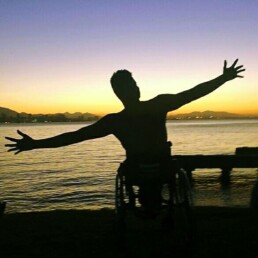 #Audiodescrição: Silhueta de pessoa de cadeira de rodas de braços abertos à beira da praia.