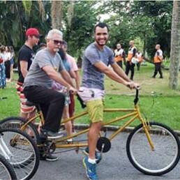 #Audiodescrição: Parque com árvores e várias pessoas, algumas de bicicleta. À frente, dois homens em uma bicicleta dupla amarela.