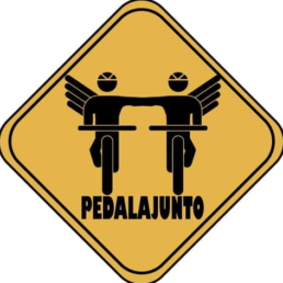 #Audiodescrição: Placa amarela, duas silhuetas em bicicletas de mãos unidas, cada uma com uma asa nas costas do lado oposto. Abaixo está escrito: Pedalajunto.