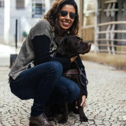 #Audiodescrição: Em uma calçada de chão de pedras cinza, Camila está agachada e abraçada com um cachorro de pelo escuro e de porte grande. Ela é branca, tem cabelo castanho longo, usa camisa preta, casaco cinza e calça jeans.
