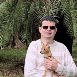 #Audiodescrição: Homem branco de cabelo preto curto. Ele usa óculos escuros, está com blusa branca e um cachorro caramelo de porte pequeno no colo. Atrás deles, uma arvore semelhante a uma palmeira e um gramado.