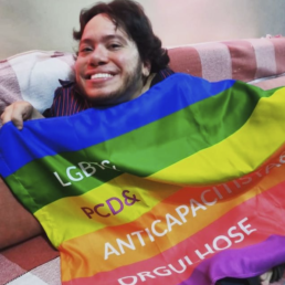 #Audiodescrição: Homem branco de cabelo e barba castanhos. Ele segura uma bandeira frente ao corpo com as cores do arco-íris escrito: LGBT & PCD & Anticapacitista % Orgulhose. Cada uma das palavras está em uma das cores da bandeira.