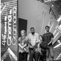 #Audiodescrição: Foto em preto e branco. Em um palco 4 pessoas, dois homens e duas mulheres. Os dois homens estão de pé ao fundo. À frente uma mulher de pé segurando um livro e outra de cadeira de rodas com um microfone em mãos.