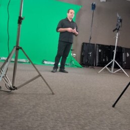 #Audiodescrição: Homem branco de cabelo preto curto, usa camisa e calça pretas. Está de pé em frente a um tecido chroma key verde, ao redor, refletores e câmeras em tripés.